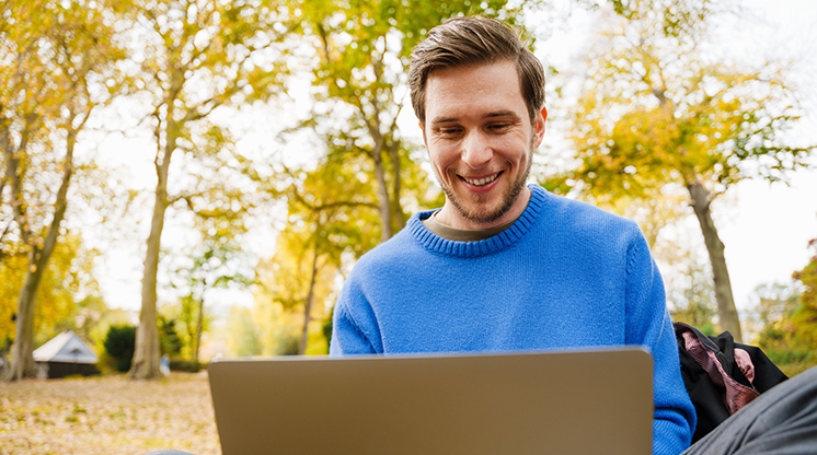 Un hombre sonriente con un suéter azul brillante se sienta afuera en su computadora